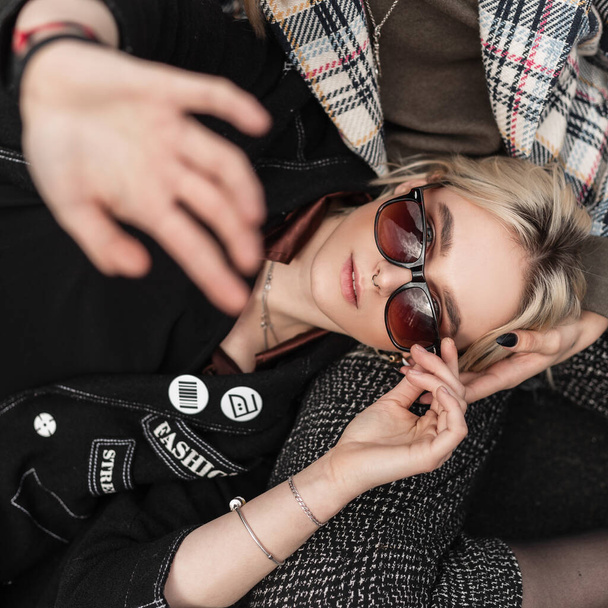 Fashion model stylish young girl in vintage black clothes in sunglasses lies and covers camera with hand. Cool trendy girlfriends resting on pavement. Youth street style. - 写真・画像