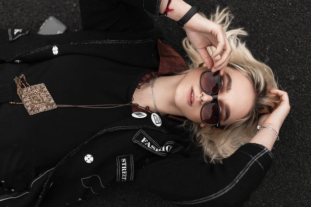 Vrij sexy mooie jonge vrouw in stijlvolle zwarte blazer in trendy zonnebril rust op straat op zomerdag op asfalt. Prachtig modern meisjesmodel ontspant in de stad. Jeugd casual mode. Bovenaanzicht. - Foto, afbeelding
