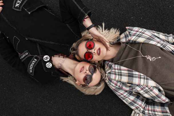 Милые привлекательные лесбиянки в повседневной модной одежде в молодежных стильных очках отдыхают на дороге на улице. Модные блондинки в винтажной одежде лежат на тротуаре в городе. Вид сверху. - Фото, изображение