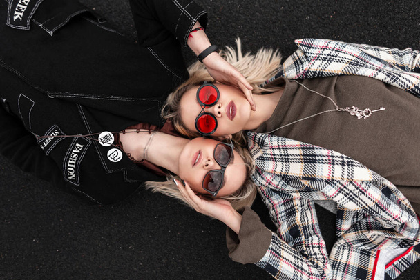 Atractivas mujeres jóvenes con estilo en gafas de moda en ropa de moda juvenil descansando en la carretera. Retrato de dos hermosas novias rubias en la calle. Vista desde arriba. - Foto, imagen