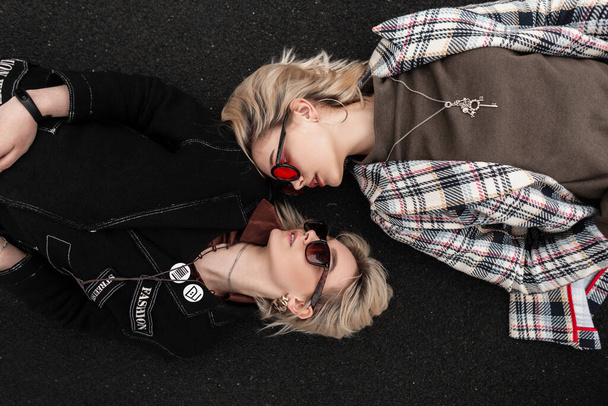 Fashionable attractive women in vintage stylish clothes in trendy sunglasses relax on asphalt and look at each other. Modern girlfriends relax on road in city. - Foto, afbeelding