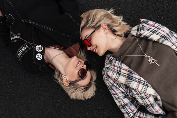 Positive beautiful woman with cute smile in black clothes with sunglasses smiles and looks at cheerful girlfriend in vintage clothes that lies nearby. Lovely girlfriends  resting on road in street. - Photo, Image