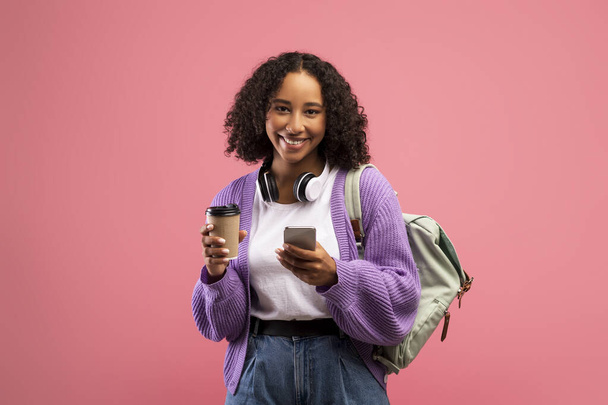 Χαρούμενη μαύρη γυναίκα με σακίδιο και ακουστικά πίνοντας καφέ, χρησιμοποιώντας το κινητό τηλέφωνο, τα μηνύματα σε απευθείας σύνδεση - Φωτογραφία, εικόνα
