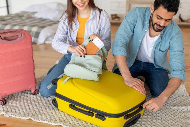 Junge Eheleute packen Kleidung in Koffer und Frau steckt Reisepass mit Fahrkarte in Tasche und bereitet sich auf Urlaub vor - Foto, Bild