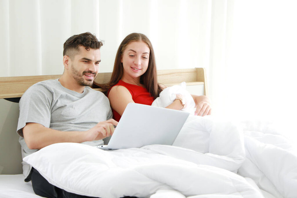 Jong Kaukasisch echtpaar houden van ontspannen en glimlachen met laptop in sociale media voor online winkelen in de slaapkamer. Technologie voor getrouwde familie levensstijl op vakantie Concept. - Foto, afbeelding