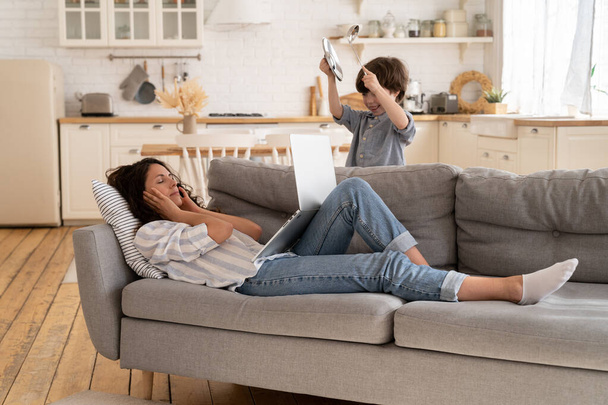 Cansado mãe estressada para se concentrar no trabalho deitado no sofá com laptop como pequeno menino rumble utensílios de cozinha - Foto, Imagem