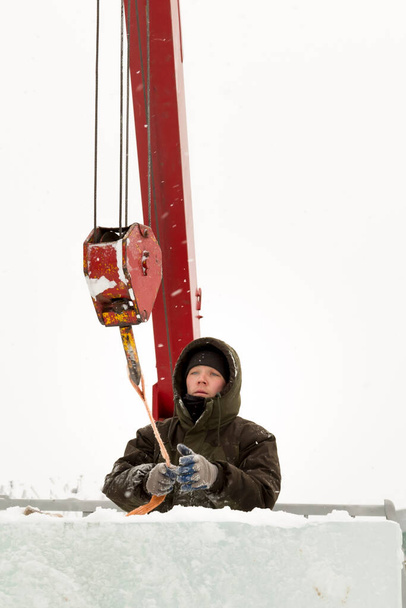 Portrait of a worker in a green jacket with a hood at the hook of a truck crane loading ice blocks - Photo, Image