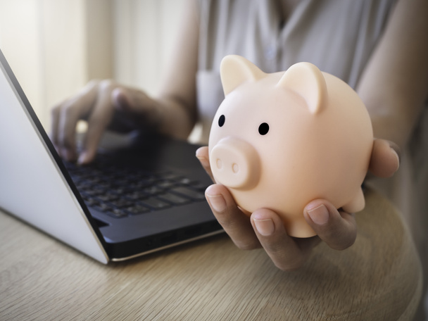 Money box on hand. Hand holding the pink piggy bank while using laptop computer on desk. Saving money wealth and financial concept, Business, online banking, finance, investment, financial planning. - Foto, Imagem