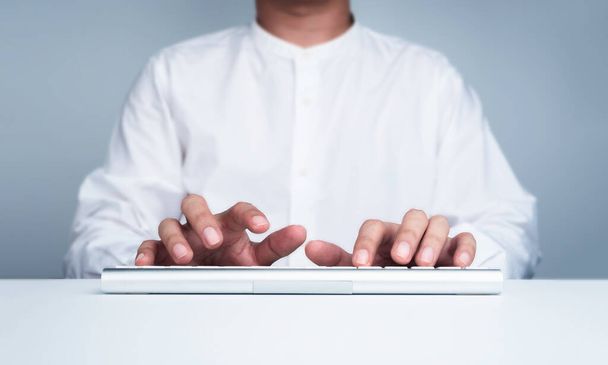 Geschäftsmannhände in weißem Hemd tippen auf einem modernen Tastaturcomputer auf einem weißen, sauberen Schreibtisch, während sie vor dem Bildschirm arbeiten, Frontansicht, minimalistischer Stil. Arbeiten mit Technologie, Online-Geschäft. - Foto, Bild