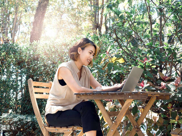 Glücklich lächelnd sitzen asiatische Frauen mit kurzen Haaren im beigefarbenen ärmellosen Hemd und arbeiten mit Laptop auf Holztisch, entspannen im Freien, umgeben von grünen Pflanzen im Garten. Arbeit überall. - Foto, Bild