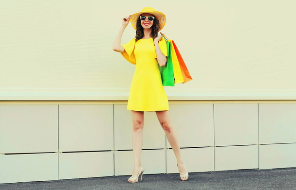 Beautiful happy smiling woman with shopping bags wearing yellow dress and summer straw hat in the city - Photo, Image