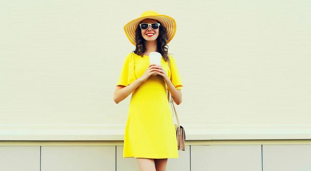 Portrait of beautiful smiling young woman with cup of coffee wearing yellow dress, summer hat on white background - Foto, imagen
