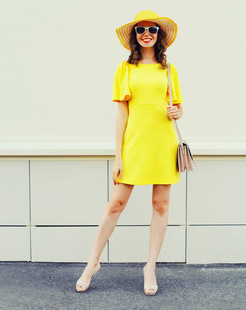 Beautiful smiling young woman in full length posing wearing yellow dress, summer straw hat, handbag on city street background - Foto, afbeelding