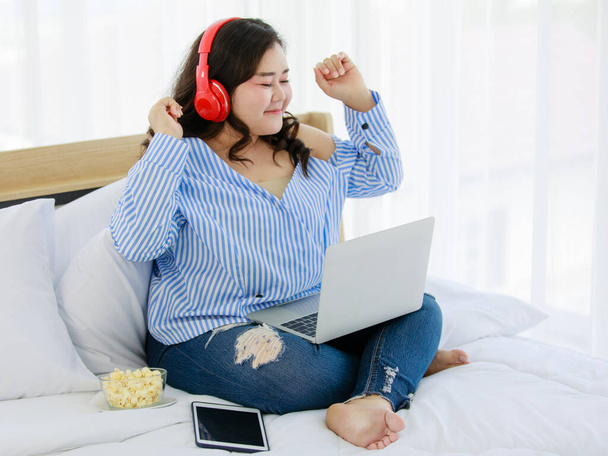 赤いヘッドフォンを身に着けているベッドルームで幸せなベッドで座って美しいぽっちゃり女性は音楽に耳を傾け、インターネットから曲をストリーミングするためにラップトップコンピュータを使用しています。女の子の幸せな生活の概念 - 写真・画像