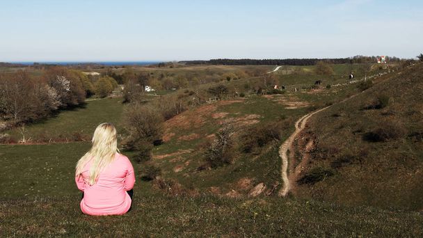 Γυναίκα με ξανθά μαλλιά κάθεται στο γρασίδι κοιτάζοντας τη θέα των λόφων, χωράφια και ανοιχτό τοπίο σε μια καλοκαιρινή μέρα. Γυναίκα που φοράει ροζ μπουφάν αντιανεμικό. Φωτογραφία: Brsarps Backar, Skne, Σουηδία. - Φωτογραφία, εικόνα