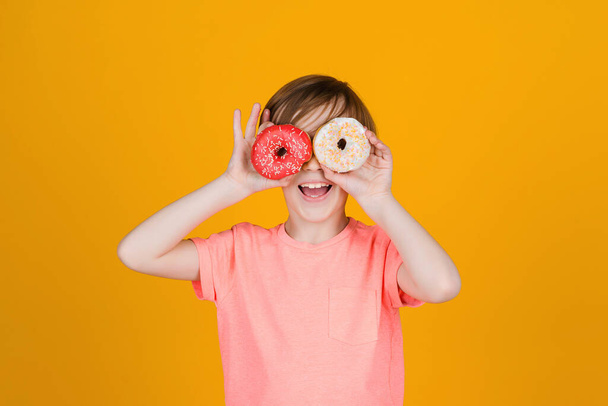 Söpö poika, jolla on donitsin silmät. Onnellinen lapsi leikki donitseilla keltaisella taustaseinällä. Poika nauttii makeisista. Syntymäpäiväjuhlat. Maukasta ruokaa lapsille. Värillisiä donitseja. Muoti, ihmiset, elämäntapa. - Valokuva, kuva