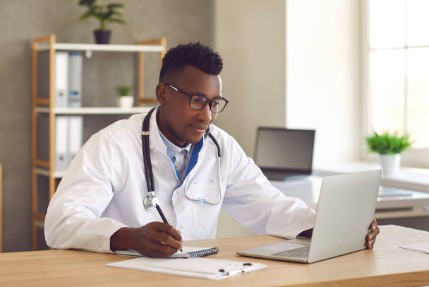 Doctor medical professional looking at laptop screen and writing notes on paper - Fotoğraf, Görsel