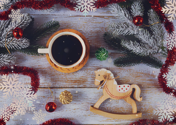 Christmas toy horse and cup of coffee on a wooden background. Festive composition with Christmas branches, cones and toys. Top view. - Photo, Image