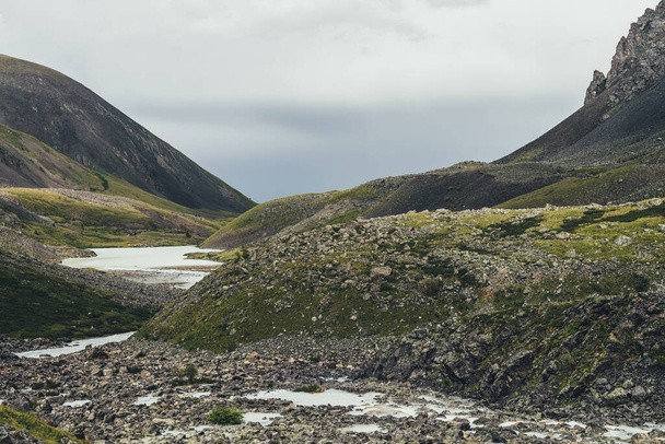 Ατμοσφαιρικό τοπίο με ορεινούς κολπίσκους και ορεινή λίμνη ανάμεσα σε βράχια σε στενή κοιλάδα με βροχερό καιρό. Bleak τοπίο με ορεινό ποτάμι και τη λίμνη μεταξύ των βράχων στην ορεινή κοιλάδα κάτω από γκρίζο συννεφιασμένο ουρανό - Φωτογραφία, εικόνα