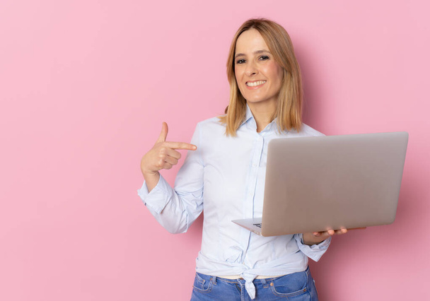 Ευτυχισμένη γυναίκα με πουκάμισο που δείχνει φορητό υπολογιστή και δείχνει σε αυτό πάνω από ροζ φόντο. - Φωτογραφία, εικόνα