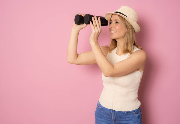 Giovane donna con cappello di paglia e guardando in lontananza con binocolo su sfondo rosa isolato - Foto, immagini