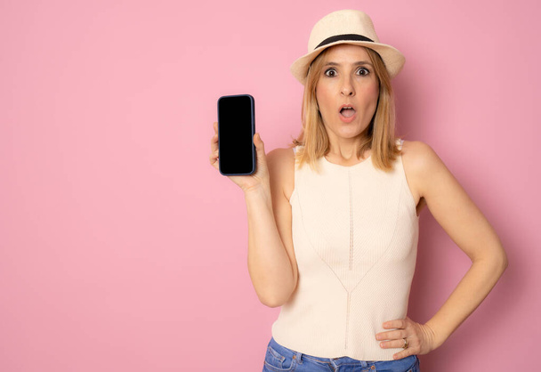 Erstaunte junge Frau mit Strohhut zeigt leeres Bildschirm-Smartphone isoliert über rosa Hintergrund - Foto, Bild
