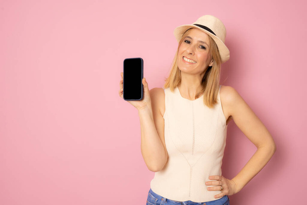 Smiling young woman in straw hat showing blank screen smartphone isolated over pink background - Photo, Image