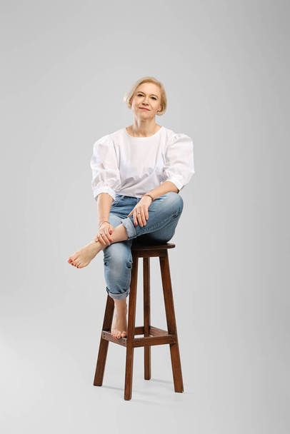 Barefoot senior woman in jeans and white blouse sitting on tall chair in studio throws one leg - Photo, image