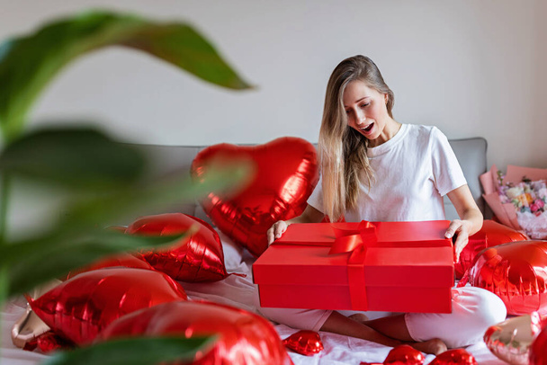 Candid lifestyle portrait of happy young caucasian woman with blonde hair and red foil heart. Millennial trendy stylish girl celebrating Valentines day 14 of February or birthday in modern apartment  - Zdjęcie, obraz