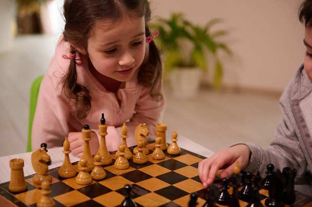 Close-up portrait of a beautiful cute little European girl playing chess game with her elder brother who picks up a chess piece and make his move. Intellectual smart development board games for kids - Foto, afbeelding