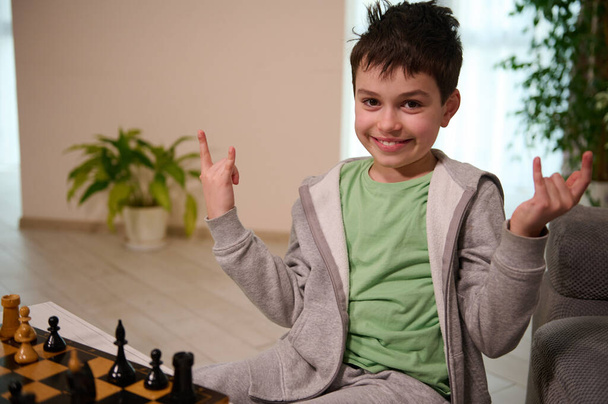 Feliz adolescente caucásico sonríe mirando a la cámara, disfrutando jugando al ajedrez en el interior del hogar - Foto, Imagen