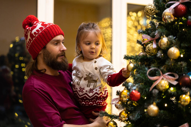 Happy dad holding a todler in Christmas clothes looking at Christmas tree . - Foto, Imagen