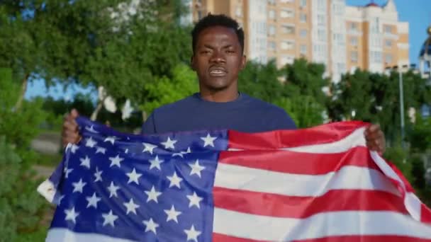 Portrait Afro-American man holding American flag looks camera says USA in summer - Materiał filmowy, wideo