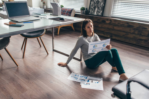 Mujer se sienta en el suelo en la oficina y mira los gráficos en la hoja - Foto, Imagen