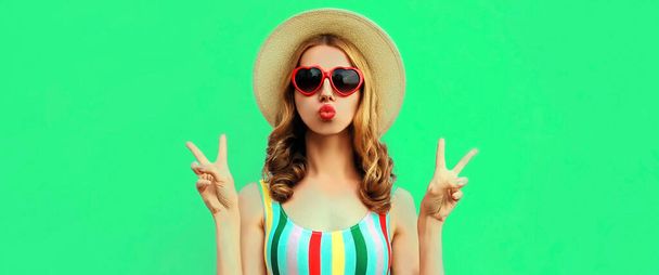 Portrait of beautiful young woman model blowing her lips and posing wearing a summer straw hat, sunglasses on green background - Photo, Image