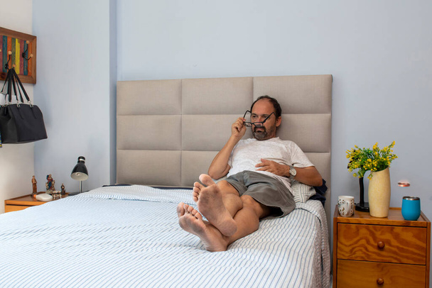 úriember ül az ágyban a hálószobában pihentető és leveszi a szemüvegét - Fotó, kép