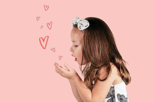 Profile of little girl in white dress and headband blowing on her hands and sending kiss and hearts on pink background - Photo, Image