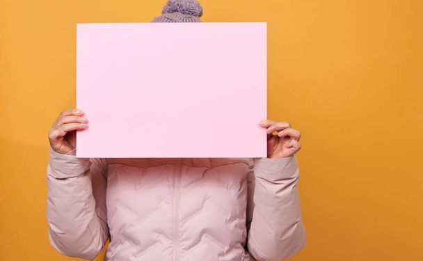 woman in pink winter jacket and hat holds blank pink sheet of paper on yellow background. Seasonal sale. Place for inscription - Foto, Imagem