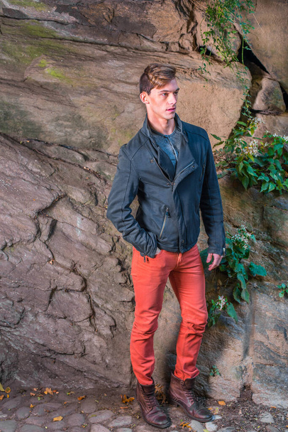 Mavi ince bir ceket, kırmızı kot pantolon, kahverengi deri ayakkabılar giyen genç bir adam kayalık bir duvarın yanında sabırla bekliyor. - Fotoğraf, Görsel