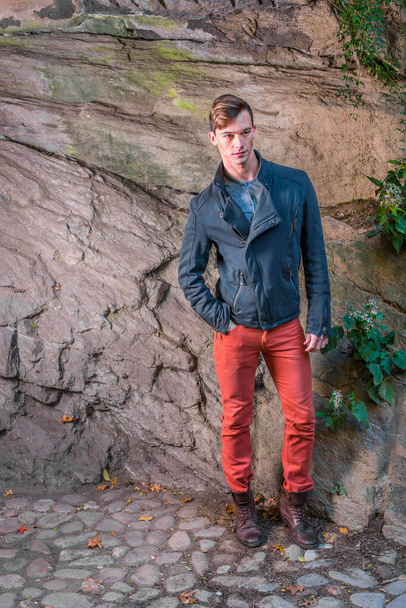 Met een blauw slim fit jasje, rode jeans, bruine leren laarsschoenen, staat een jongeman bij een rotsachtige muur, geduldig wachtend. - Foto, afbeelding