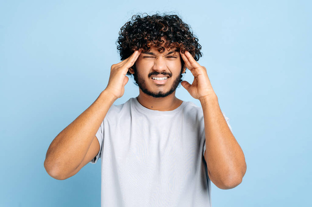 Upset unhealthy indian guy, grimacing in pain in his head, massaging his temples, experiencing a migraine, headache, overworked, in need of rest or treatment, stands on an isolated blue background - 写真・画像