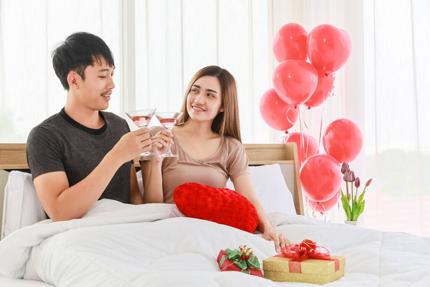 Lovely couple and Valentine's Day Concept. Smiling asian man and woman drinking sparkling wine in bed at bedroom with red balloon and beautiful gift box. - Photo, Image