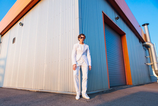 Wide angle portrait of a handsome man dressed in a white suit and round hippie sunglasses standing near the corner of a building during sunset - Fotoğraf, Görsel