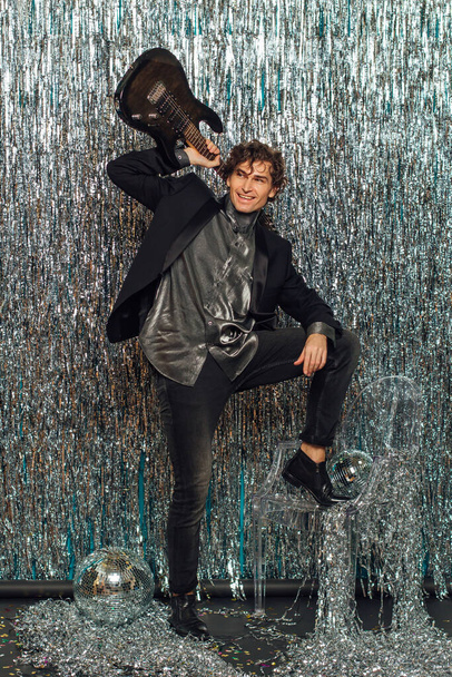 Tall attractive man holding electro guitar standing over the sparkle background with a mirror disco ball on the floor. Party concept. - Foto, afbeelding