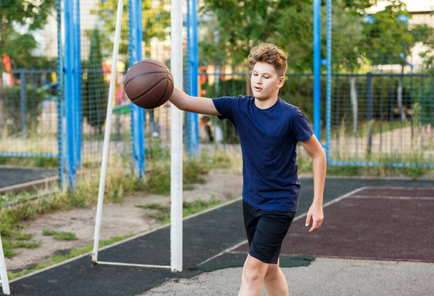 Netter lächelnder Junge im blauen T-Shirt spielt Basketball auf dem städtischen Spielplatz. Aktive Teenager genießen ein Spiel im Freien mit einem orangefarbenen Ball. Hobby, aktiver Lebensstil, Sport für Kinder, Jugendliche - Foto, Bild