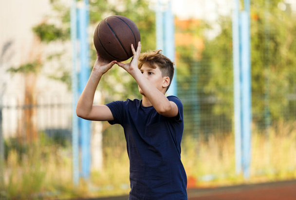 Netter lächelnder Junge im blauen T-Shirt spielt Basketball auf dem städtischen Spielplatz. Aktive Teenager genießen ein Spiel im Freien mit einem orangefarbenen Ball. Hobby, aktiver Lebensstil, Sport für Kinder, Jugendliche - Foto, Bild
