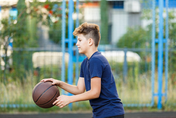 Lindo niño sonriente en una camiseta azul juega baloncesto en el patio de la ciudad. Adolescente activo disfrutando de un juego al aire libre con una bola naranja. Aficion, estilo de vida activo, deporte para niños, adolescentes - Foto, imagen