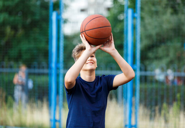 Roztomilý usmívající se chlapec v modrém tričku hraje basketbal na městském hřišti. Aktivní dospívající těší venkovní hru s oranžovou koulí. Hobby, aktivní životní styl, sport pro děti, teenageři - Fotografie, Obrázek