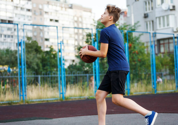 Милый улыбающийся мальчик в синей футболке играет в баскетбол на городской площадке. Активный подросток наслаждается игрой на открытом воздухе с оранжевым мячом. Хобби, активный образ жизни, спорт для детей, подростков - Фото, изображение