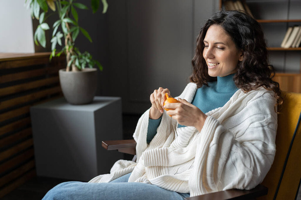 Joyeuse femme heureuse peler mandarine juteuse fraîche relaxante dans un fauteuil confortable sous couverture à la maison - Photo, image